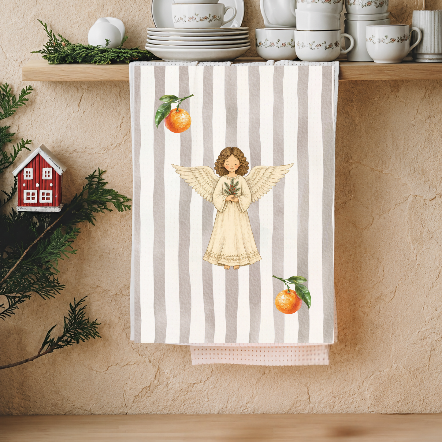 Angels and oranges Christmas tea towel hanging from a kitchen shelf of white china, with pine sprigs and a small red house