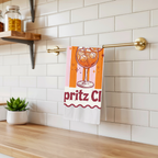 Spritz Club tea towel hanging from a brass rail against white subway tile