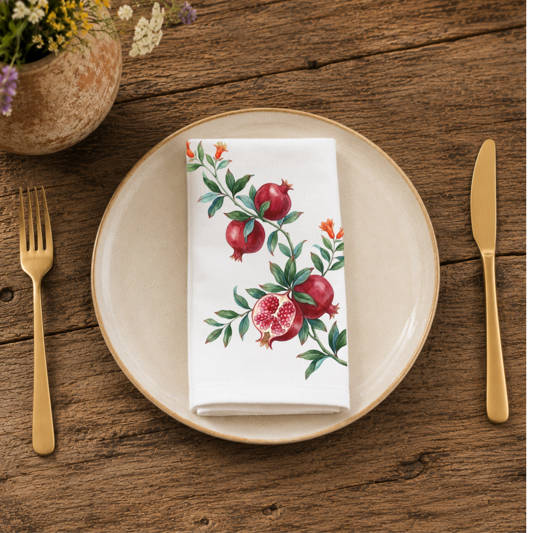 Pomegranate napkin folded on a cream plate between gold cutlery, beside a terracotta vase of Mediterranean wildflowers
