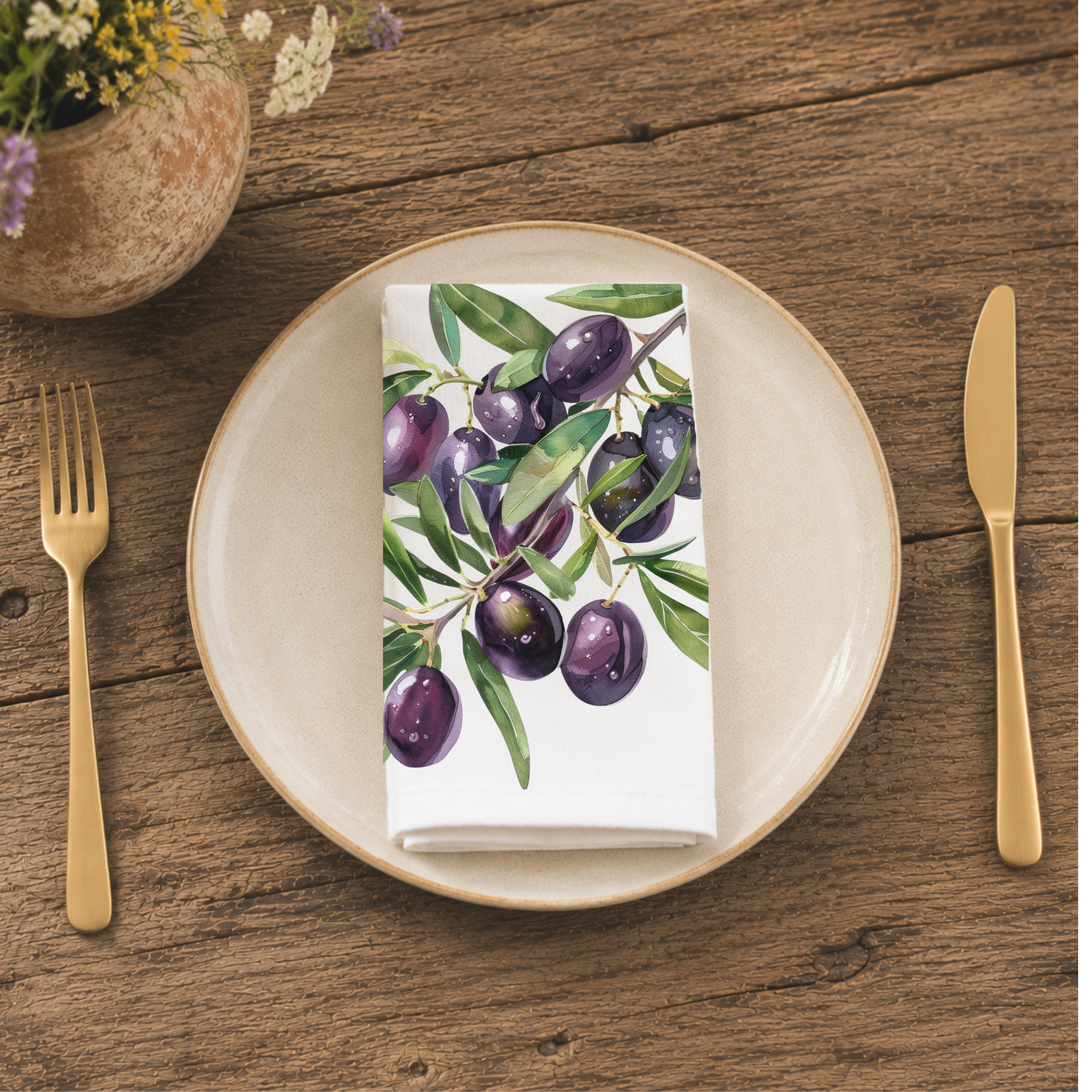 Olive Branch napkin folded on a cream plate between gold cutlery, beside a terracotta vase of Mediterranean wildflowers