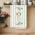 Angels and holly Christmas tea towel hanging from a kitchen shelf of white china, with pine sprigs and a small red house