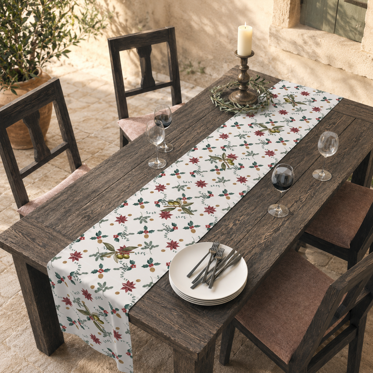 Poinsettia and olive Christmas table runner on a dark wood table in a sunlit courtyard, with white plates and red wine