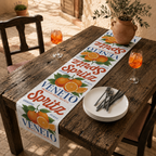 Spritz Veneto table runner on a weathered wooden table in a sunlit courtyard, with two glasses of orange spritz, stacked plates and an olive branch arrangement
