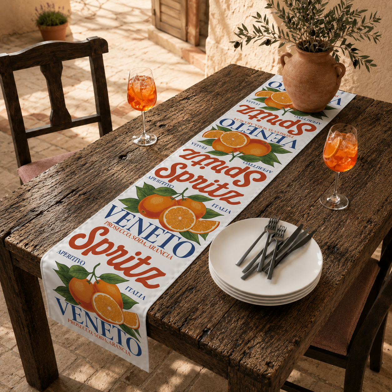 Spritz Veneto table runner on a weathered wooden table in a sunlit courtyard, with two glasses of orange spritz, stacked plates and an olive branch arrangement