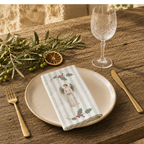 Angels and holly napkin folded on a stoneware plate in warm sunlight, with gold cutlery, a wine glass and an olive branch