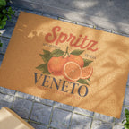 Spritz Veneto coir doormat on blue-grey stone paving in warm sunlight, with the Spritz Veneto label print and oranges clearly visible on the coir