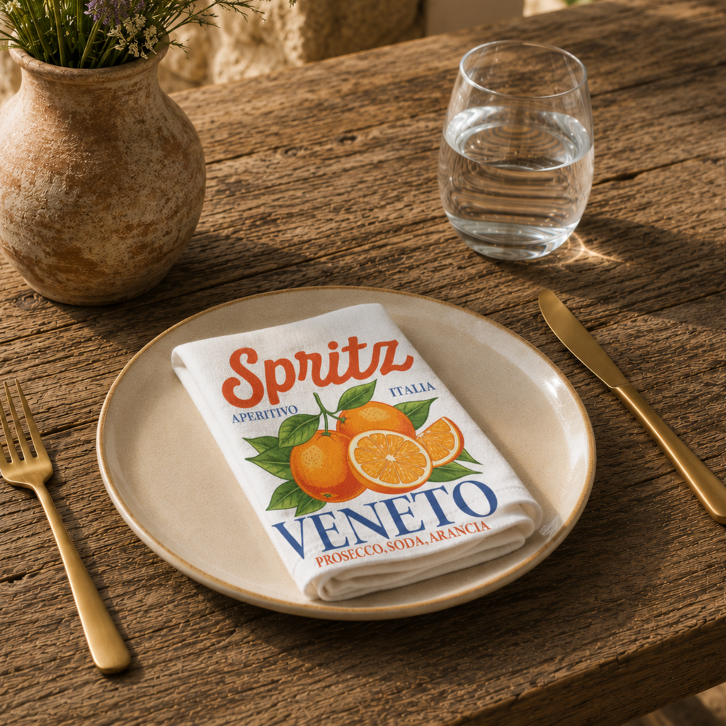 Spritz Veneto cloth napkin folded on a stoneware plate in warm sunlight, with gold cutlery, a glass of water and a lavender arrangement