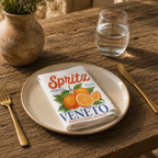 Spritz Veneto cloth napkin folded on a stoneware plate in warm sunlight, with gold cutlery, a glass of water and a lavender arrangement