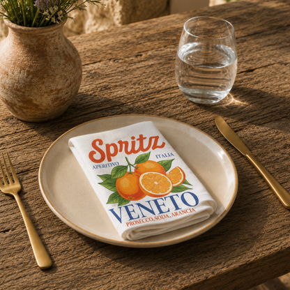 Spritz Veneto cloth napkin folded on a stoneware plate in warm sunlight, with gold cutlery, a glass of water and a lavender arrangement