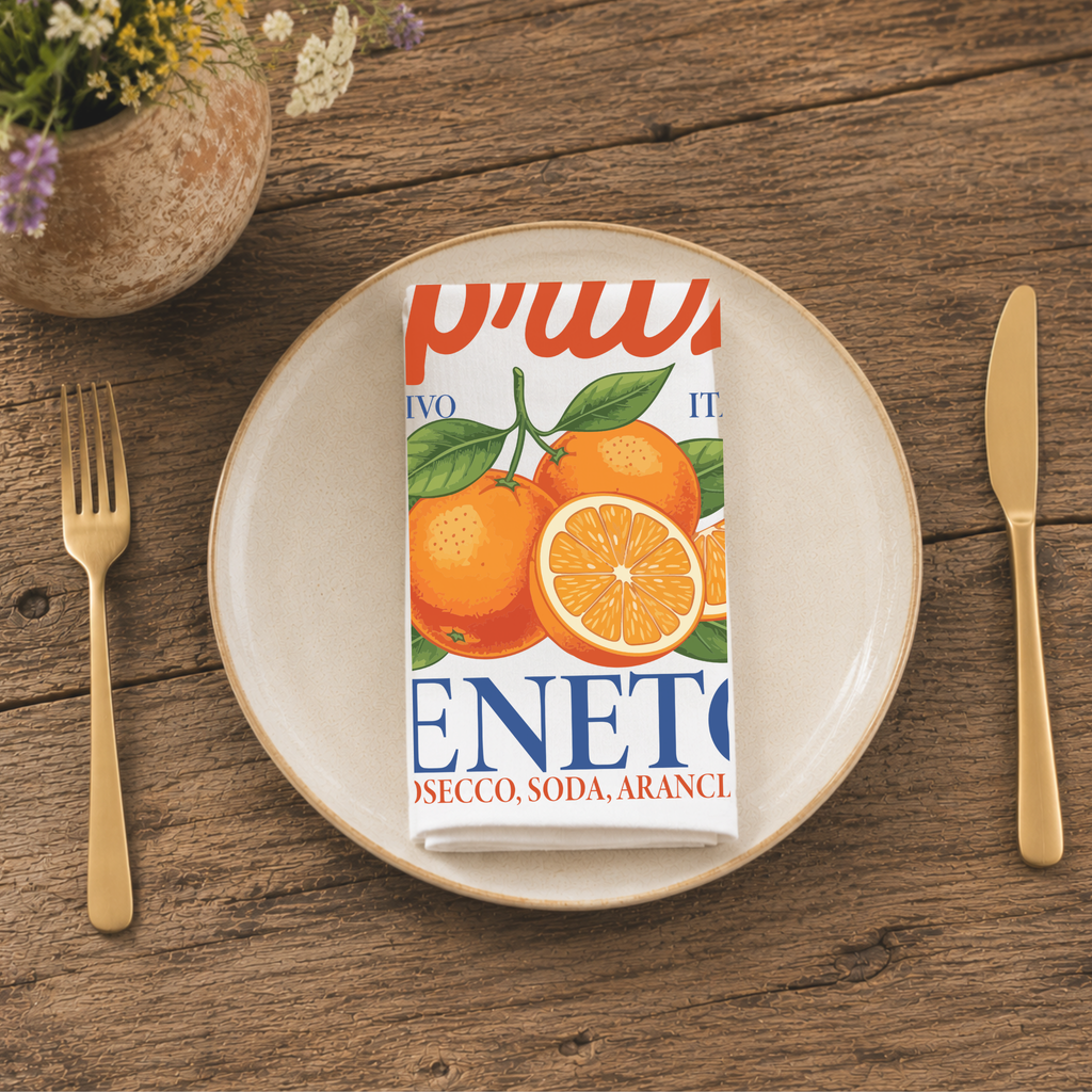 Spritz Veneto cloth napkin folded on a stoneware plate, seen from above with gold cutlery and a pot of wildflowers on a wooden table
