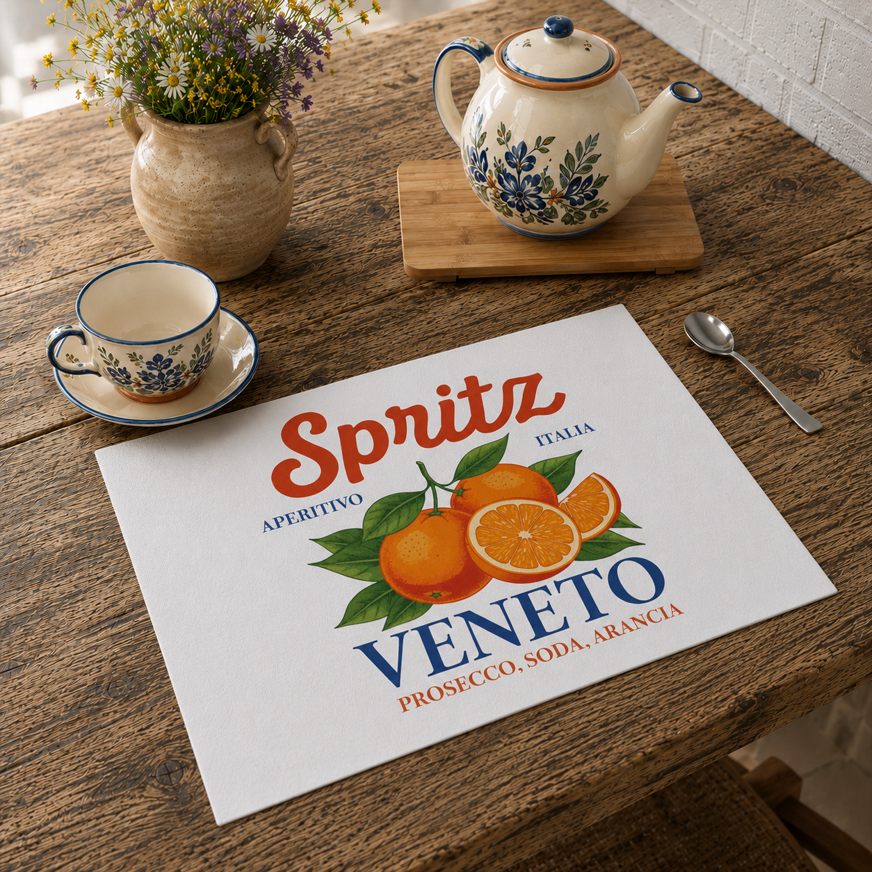 Spritz Veneto placemat on a rustic wooden table with a ceramic cup and saucer, a floral teapot and a vase of wildflowers