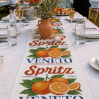 Spritz Veneto table runner on a long white-clothed table at an outdoor gathering, with champagne flutes, a lit candle and an olive branch in a terracotta vase