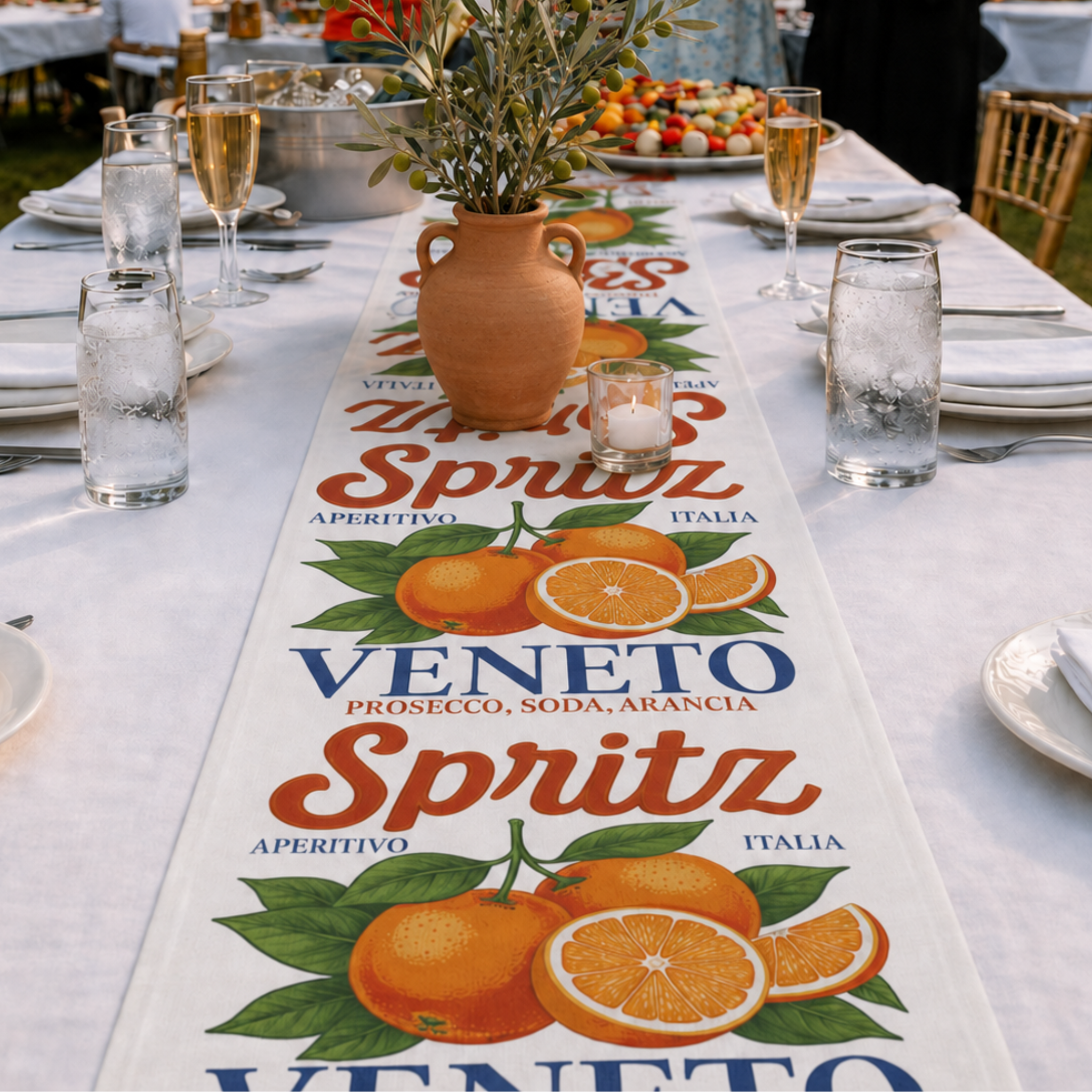 Spritz Veneto table runner on a long white-clothed table at an outdoor gathering, with champagne flutes, a lit candle and an olive branch in a terracotta vase
