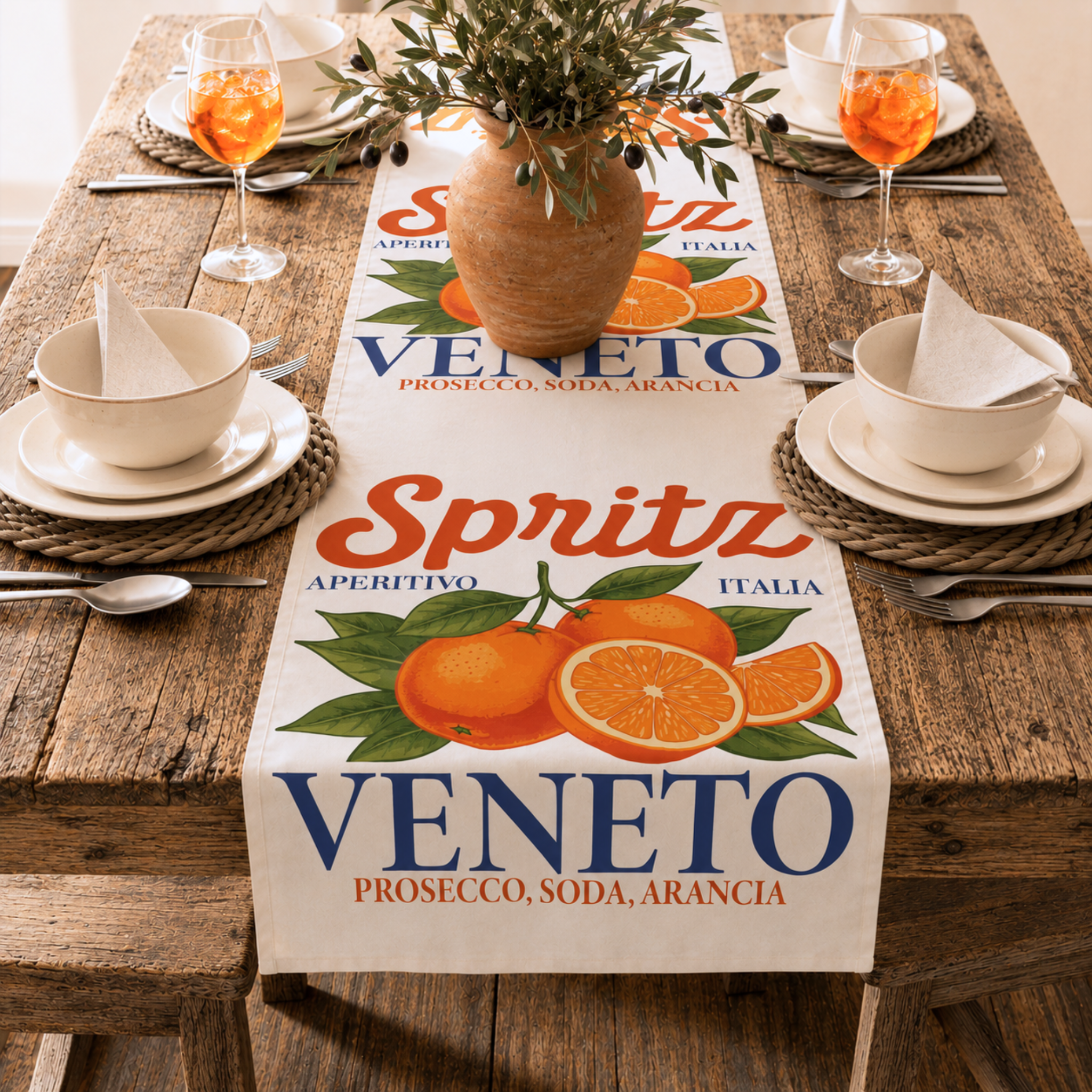 Spritz Veneto table runner running the length of a rustic wooden table set for four, with woven placemats, cream bowls and an olive branch arrangement