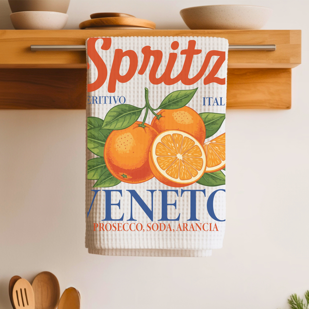 Spritz Veneto tea towel draped over a wooden shelf rail, with ceramic bowls on the shelf above and wooden utensils below