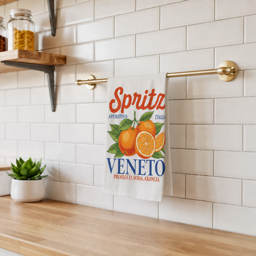 Spritz Veneto tea towel on a brass rail against white subway tile, beside a wooden shelf and a small potted succulent